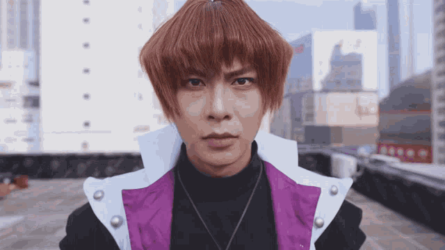 a man wearing a purple vest and a black turtleneck is looking at the camera