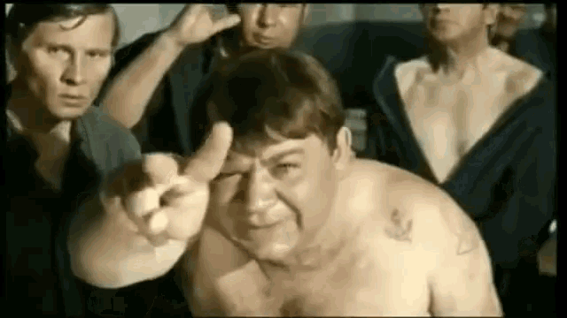 a group of men are standing around a shirtless man who is pointing at them .