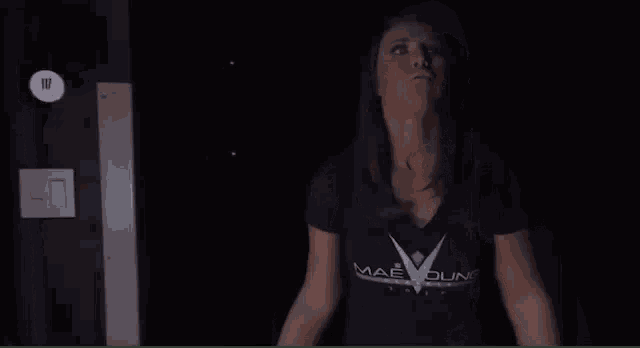 a woman in a mae young t-shirt is standing in the dark .