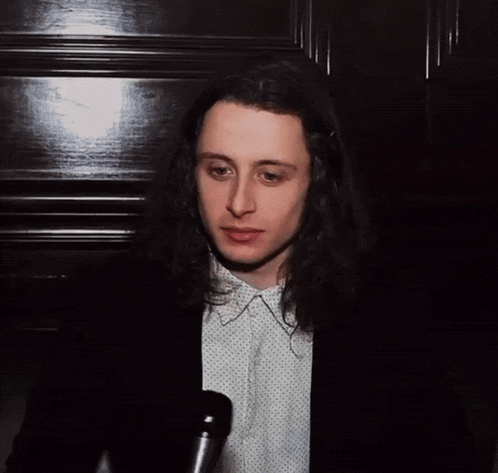 a man with long hair is wearing a black jacket and a white polka dot shirt