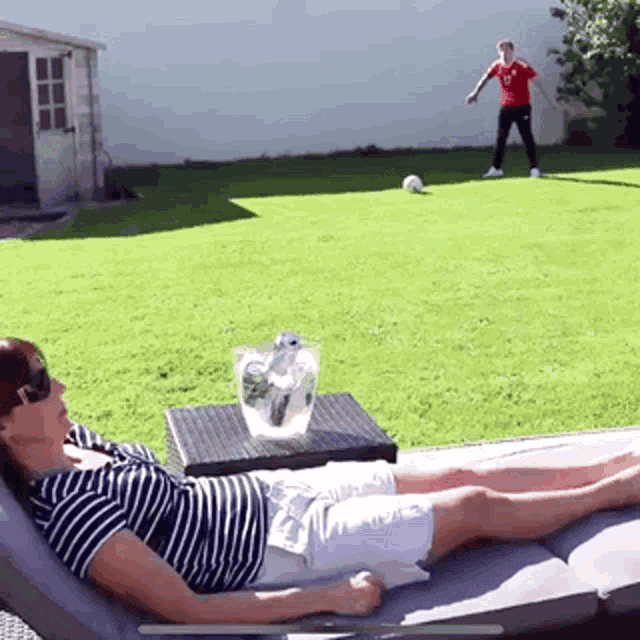 a man in a red shirt is kicking a soccer ball while a woman sits on a lounge chair