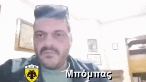 a man with a mustache is taking a selfie while wearing sunglasses and a greek shirt .