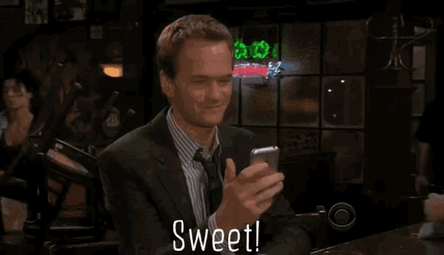a man in a suit and tie is holding a cell phone in front of a neon sign that says " sweet "