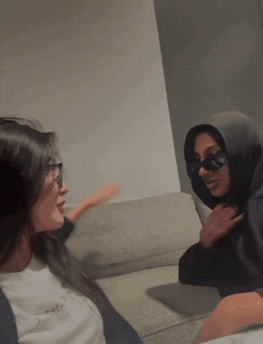 two women sitting on a couch with one wearing a hoodie and the other wearing sunglasses