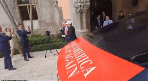 a man in a white hat stands in front of a red sign that says " make america great again "