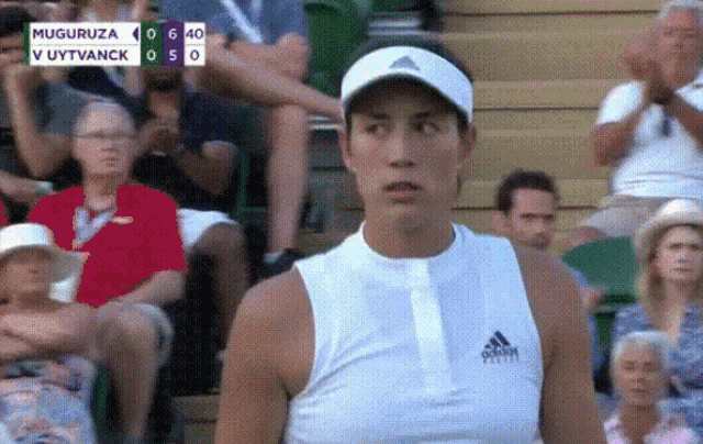 a woman wearing a white adidas shirt stands in front of a crowd watching a tennis match