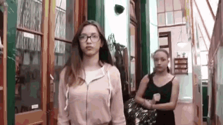two girls are walking down a hallway in a building . one of the girls is wearing glasses .