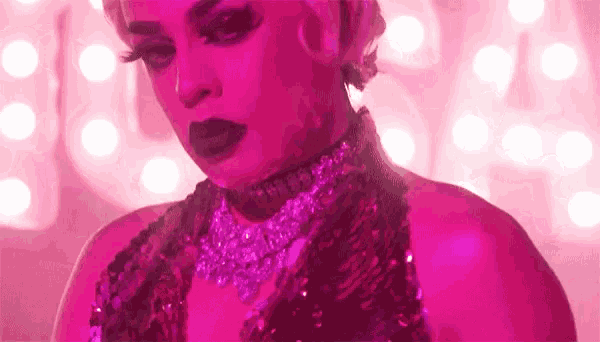 a drag queen is standing in front of a pink background wearing a choker and a necklace .