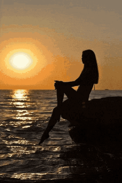 a silhouette of a woman sitting on a rock near the ocean at sunset