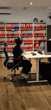 a man sits at a desk with a laptop and binders in the background