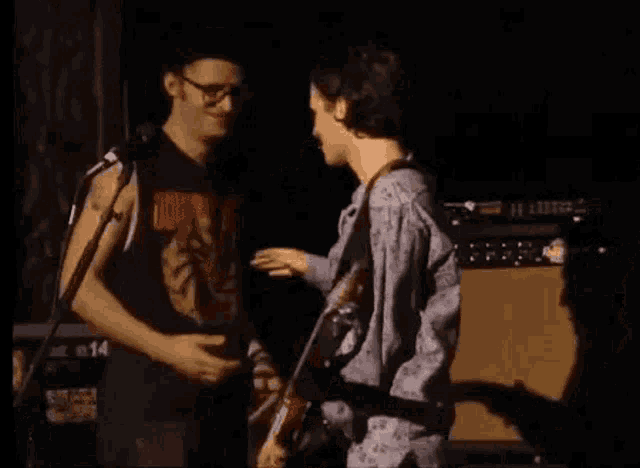 a man is playing a bass guitar on a stage while another man stands behind him .