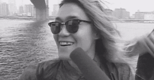 a woman wearing sunglasses is smiling in a black and white photo with her hair blowing in the wind .