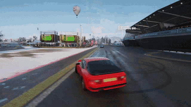 a red car is driving down a snowy road in front of a stadium with a hot air balloon flying in the sky
