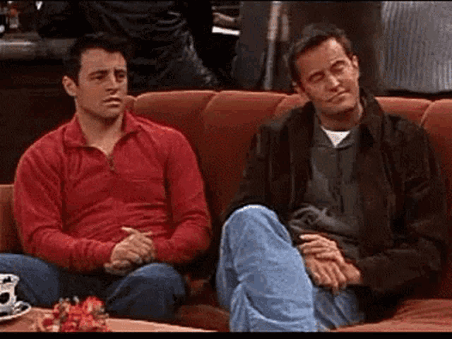 two men are sitting on a couch with their legs crossed . one of the men is wearing a red shirt .
