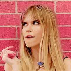 a woman with long blonde hair is standing in front of a pink brick wall and making a funny face .