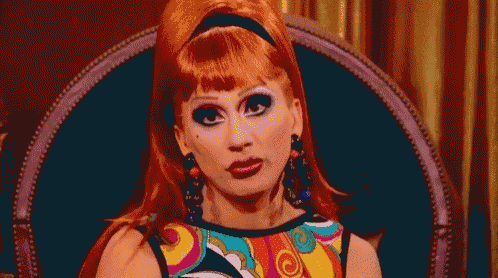 a drag queen with red hair is sitting in a chair wearing a colorful dress and earrings .