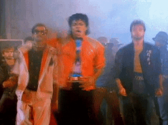 a group of men are standing next to each other in a dark room . one of the men is wearing an orange jacket .