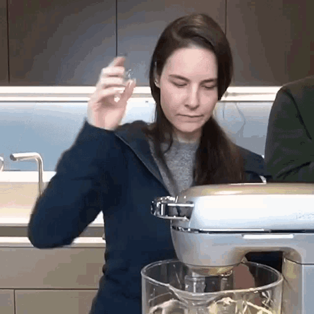 a woman in a blue jacket is using a mixer in a kitchen while a man in a green shirt looks on