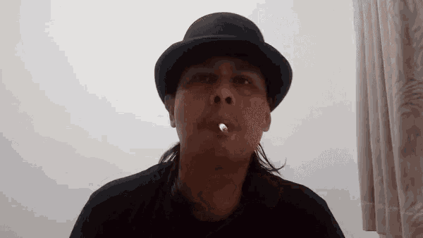 a man wearing a bowler hat is smoking a cigarette in front of a white wall .