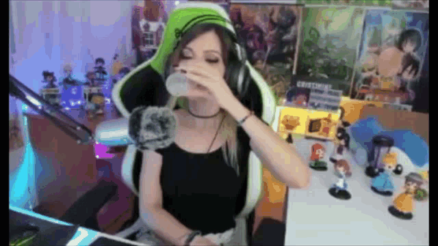 a woman wearing headphones and a green hat is drinking from a cup while sitting in a gaming chair .