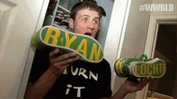 a man wearing a black shirt that says turn it is holding up a pair of shoes with the name ryan on them