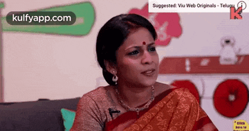 a woman in a red saree is sitting on a couch with a microphone in her mouth .