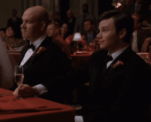 two men in tuxedos are sitting at a table with a glass of wine