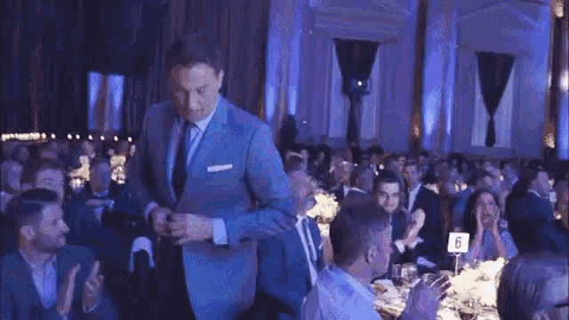 a man in a suit and tie is standing in front of a crowd of people sitting at tables .