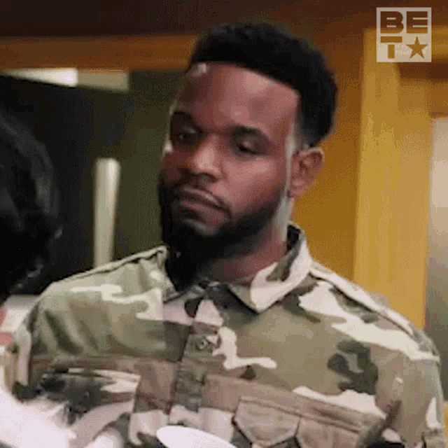 a man with a beard wearing a camouflage jacket is standing in a kitchen .