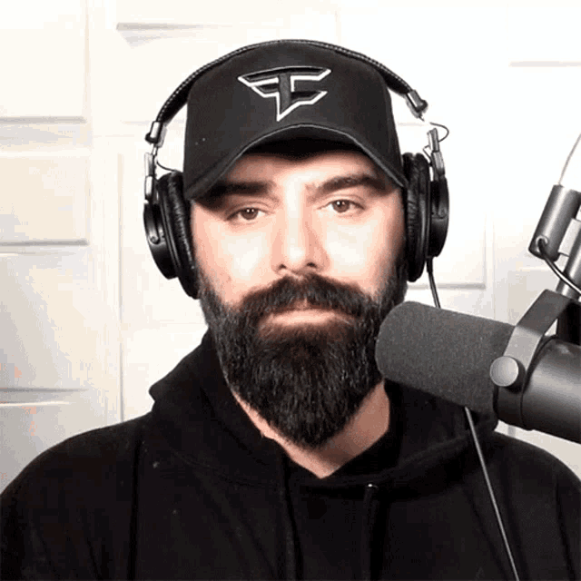 a man with a beard wearing headphones and a hat with the letter f on it stands in front of a microphone