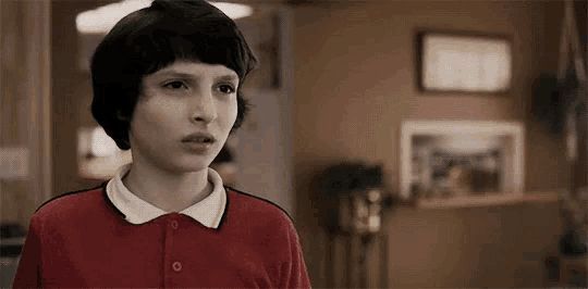 a young boy in a red shirt and white collar is standing in a room and looking at the camera .