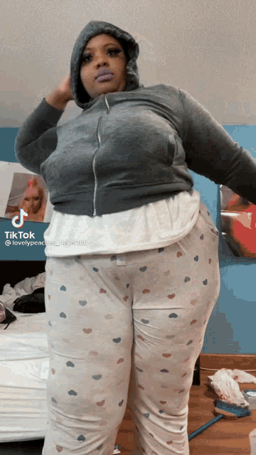 a woman wearing a grey hoodie and white pajama pants with hearts on them is standing in front of a bed