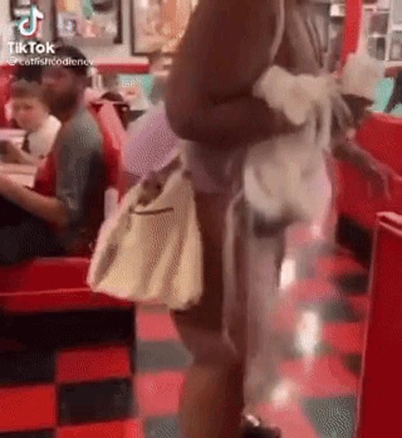a woman is standing in a diner holding a purse while a man sits at a table .