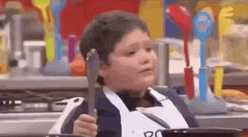 a young boy in an apron is holding a spatula and a pan .