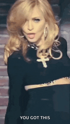 a woman in a black dress with a cross necklace is standing in front of a brick wall and says you got this .