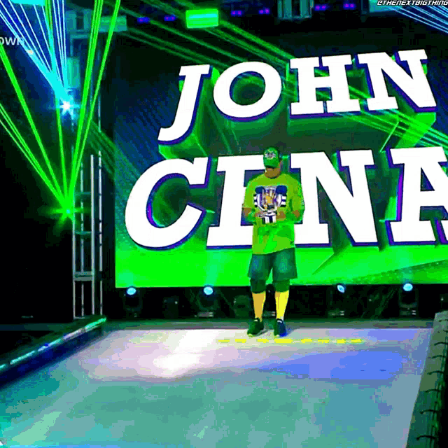a man in a green shirt stands on a stage in front of a large screen that says john cena