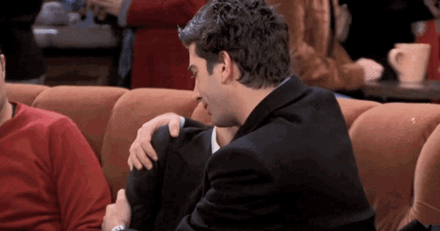 a man in a suit holds another man 's shoulder while sitting on a couch