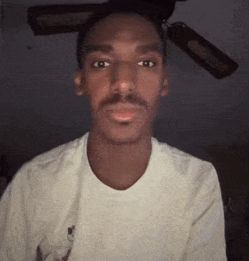a man with a mustache is wearing a white shirt and a ceiling fan in the background