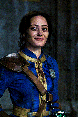 a woman wearing a blue jacket with a brown strap around her shoulder looks at the camera
