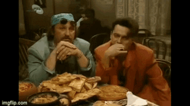 two men are sitting at a table with a bunch of food on it .
