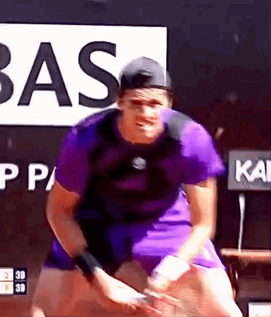a man in a purple shirt is swinging a tennis racket in front of a sign that says bas