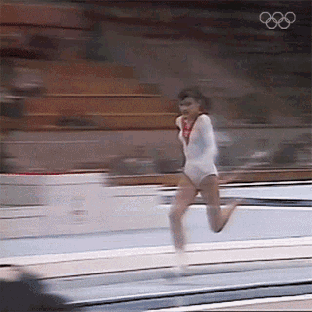 a woman in a white leotard is jumping on a trampoline with the olympic rings behind her
