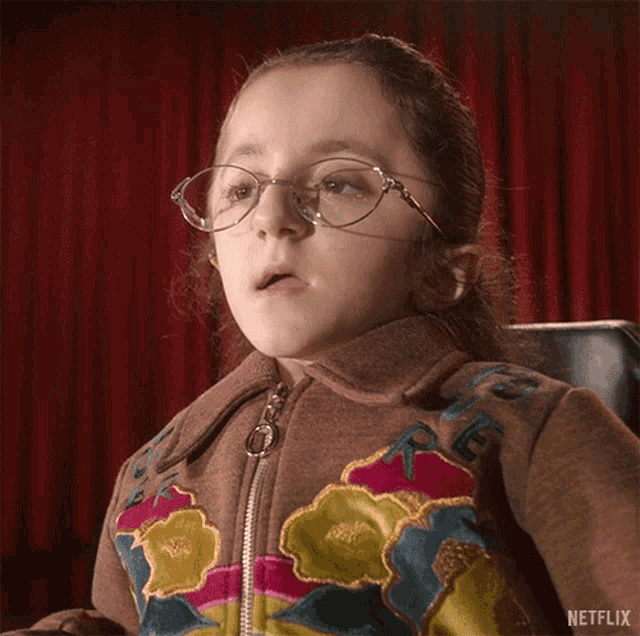 a little girl wearing glasses and a jacket with flowers on it is sitting in a chair .