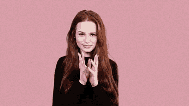 a woman with long red hair is making a heart shape with her hands against a pink background .