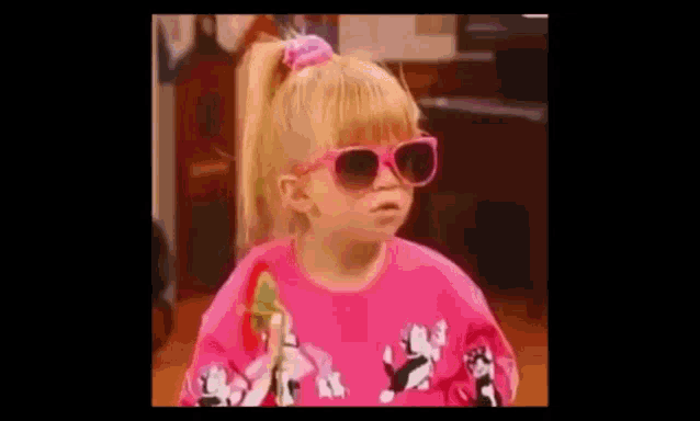 a little girl wearing pink sunglasses and a pink sweater is holding a lollipop .