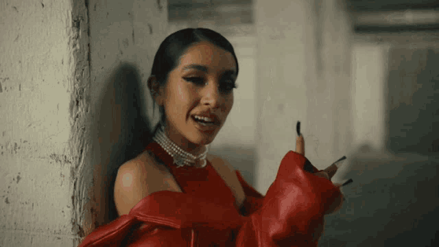 a woman leaning against a wall wearing a red jacket and a pearl necklace