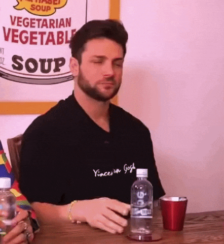 a man with a beard is sitting at a table with a bottle of water and a cup .