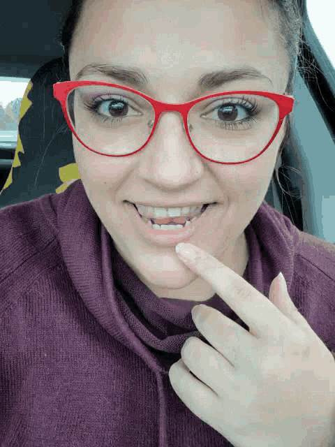 a woman wearing red glasses and a purple sweater is pointing to her mouth .