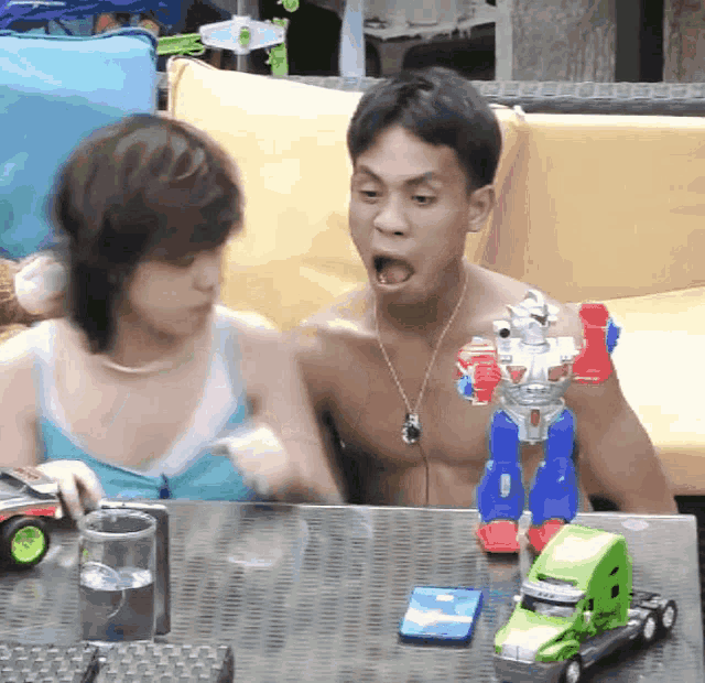 a shirtless man sits at a table with a toy robot