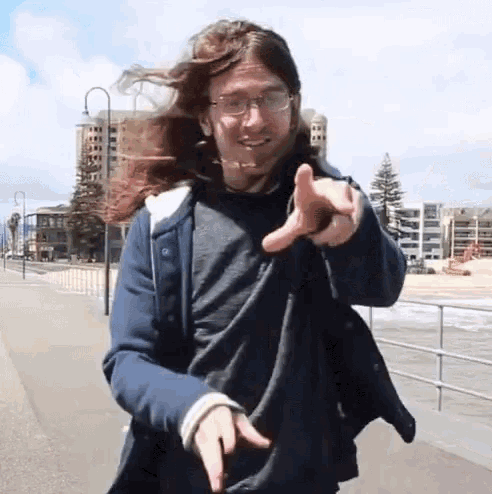 a man with long hair wearing glasses and a blue jacket is pointing at the camera